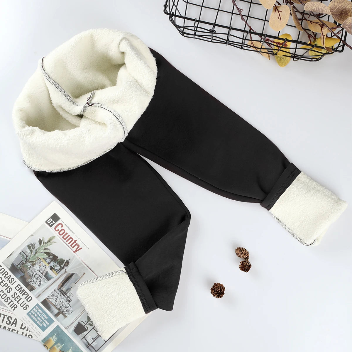 Women's velvet winter leggings in black, flat lay with waistband and ankle cuffs rolled showing thick cream fleece interior lining, styled with magazine and pine cones, top view