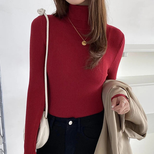 Woman wearing burgundy red cashmere blend ribbed turtleneck sweater with navy pants and gold necklace, holding beige jacket, front view