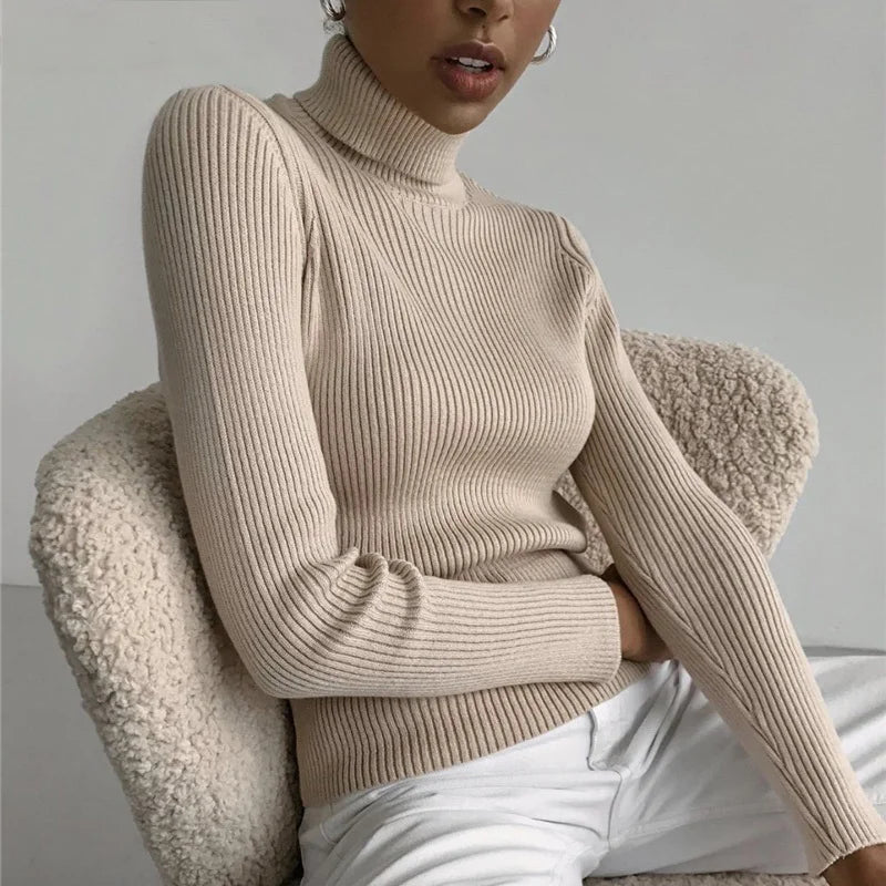 Woman wearing cream cashmere blend ribbed turtleneck sweater seated on white armchair with white pants, lifestyle front view