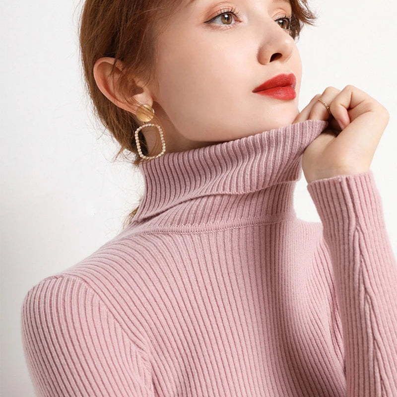 Woman wearing dusty pink cashmere blend ribbed turtleneck sweater with gold square earrings, hand at collar, front view