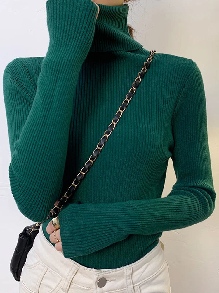 Woman wearing emerald green cashmere blend ribbed turtleneck sweater with white pants and black chain bag, front view