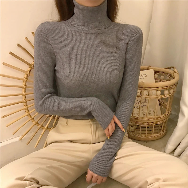 Woman wearing light gray cashmere blend ribbed turtleneck sweater with beige pants seated on rattan chair, front view