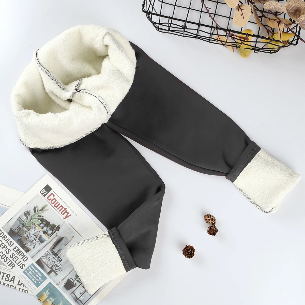 Women's velvet winter leggings in black, flat lay with waistband and ankle cuffs rolled showing thick cream fleece interior lining, styled with magazine and pine cones