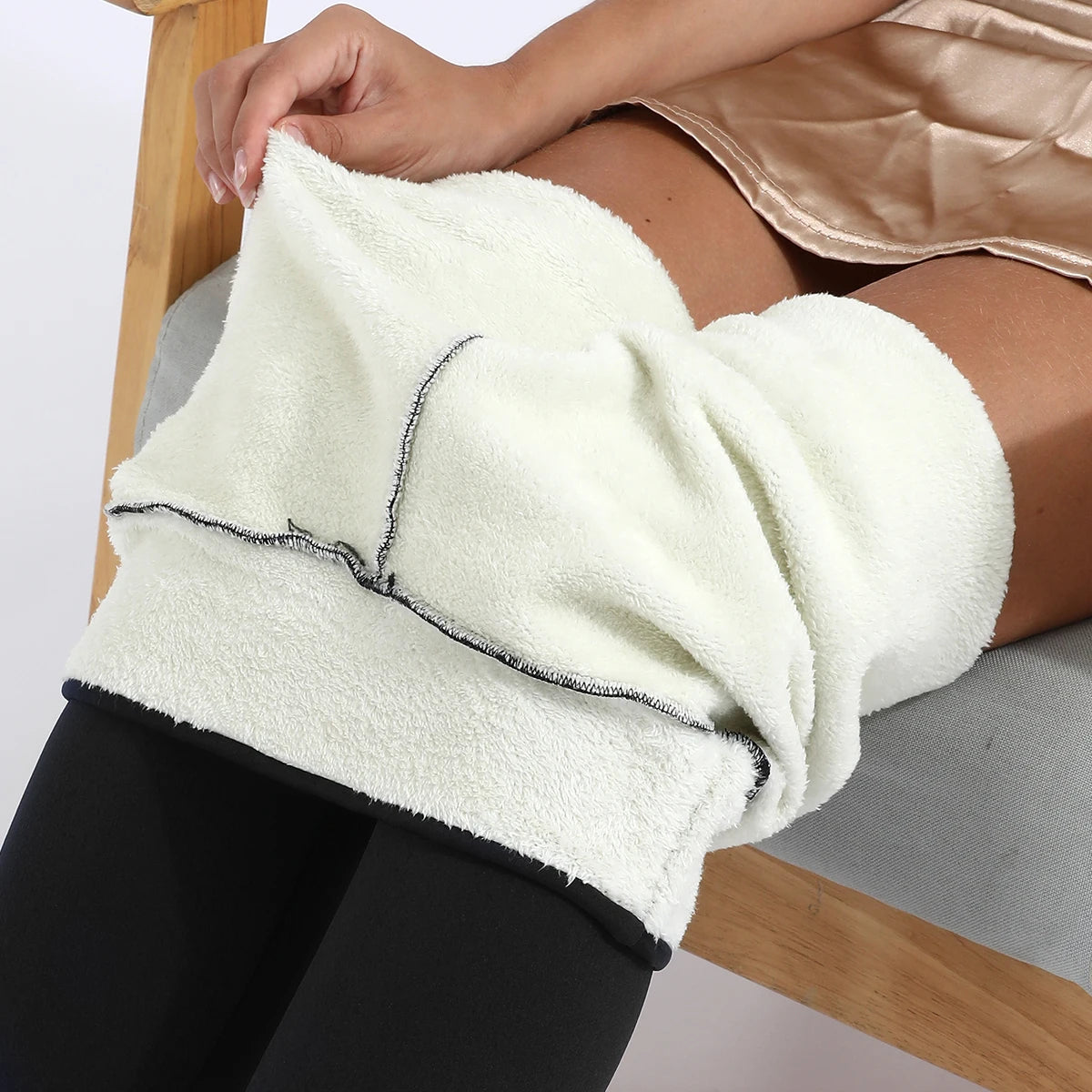 Women's high waist velvet winter leggings in black, close-up of waistband rolled down showing thick plush cream fleece interior lining, held by model seated on chair
