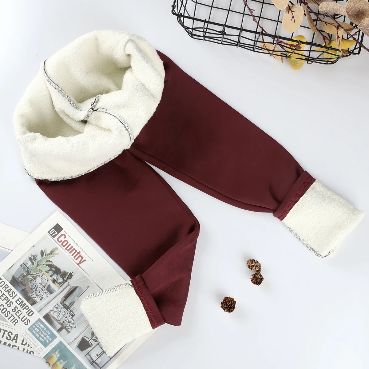 Women's velvet winter leggings in burgundy, flat lay with waistband and ankle cuffs rolled showing thick cream fleece interior lining, styled with magazine and pine cones