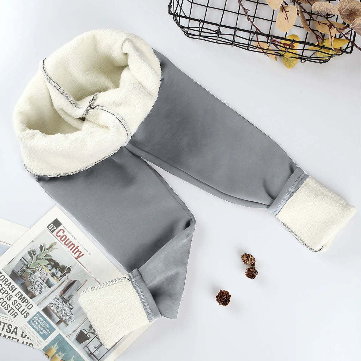 Women's velvet winter leggings in gray, flat lay with waistband and ankle cuffs rolled showing thick cream fleece interior lining, styled with magazine and pine cones