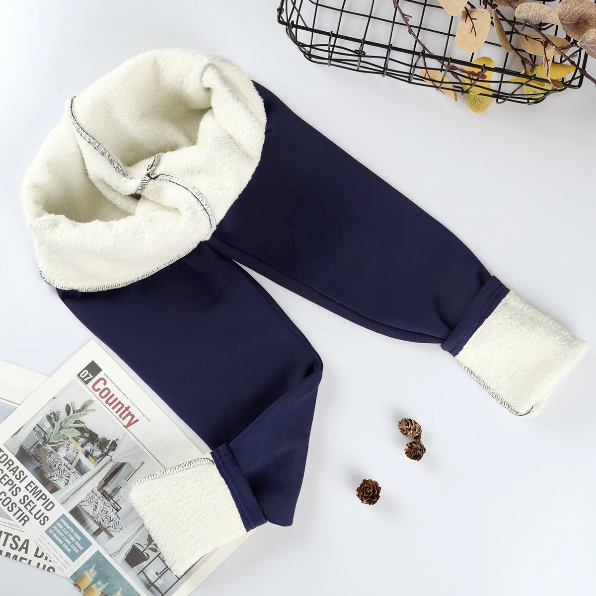 Women's velvet winter leggings in navy blue, flat lay with waistband and ankle cuffs rolled showing thick cream fleece interior lining, styled with magazine and pine cones