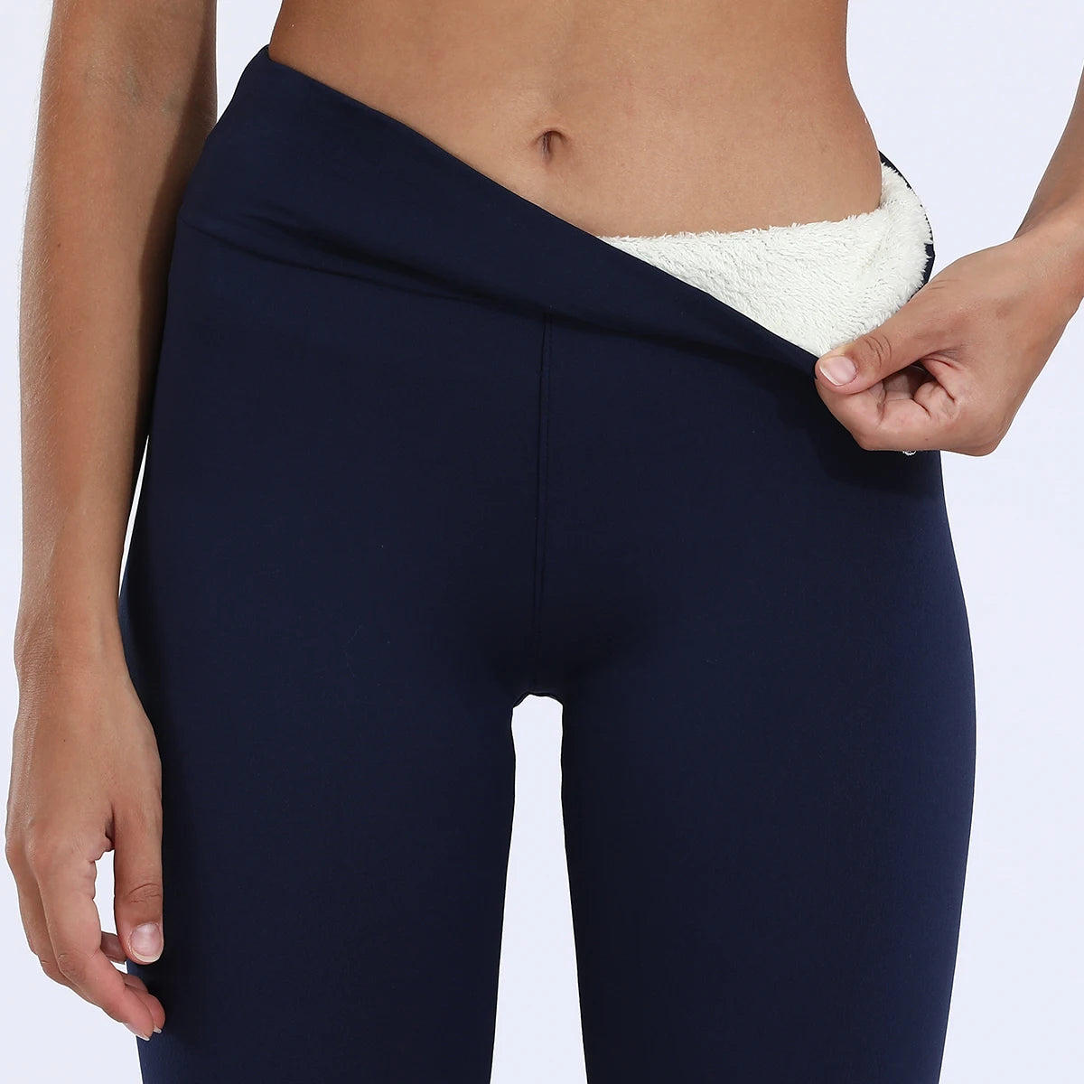 Women's high waist velvet winter leggings in navy blue, front close-up of waistband pulled down by model showing thick cream fleece interior lining detail