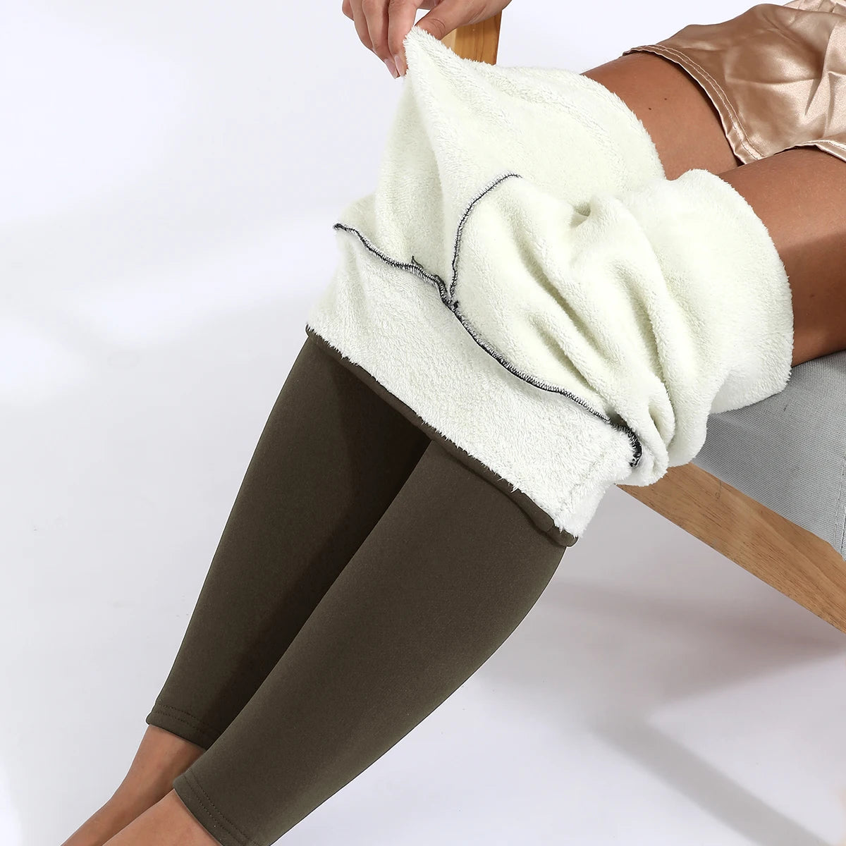 Women's high waist velvet winter leggings in olive green, close-up of waistband rolled down showing thick plush cream fleece interior lining, model seated on wooden chair